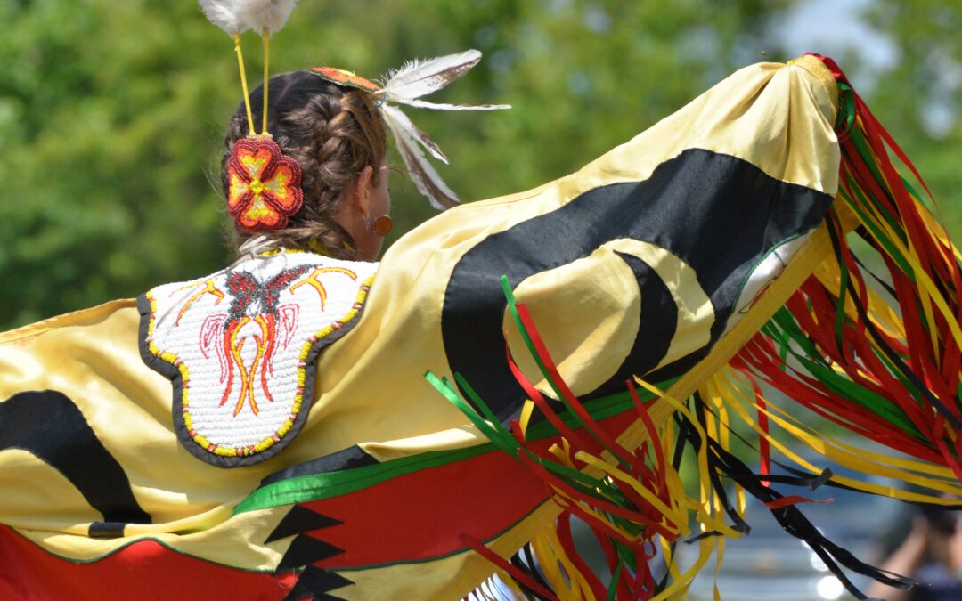 An Indigenous dancer, dancing outside with greenery from trees in the background.